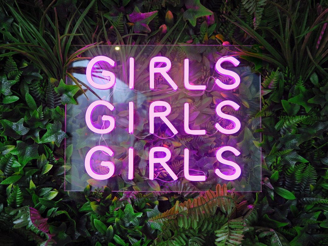 A pink neon sign saying 'Girls Girls Girls' against a garden backdrop.